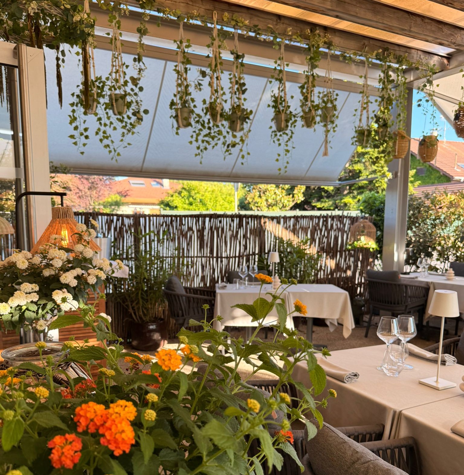 Vue large de la terrasse de l'Auberge d'Arthaz avec lantanas oranges et blanches au premier plan, tables dressées et herbes séchées suspendues