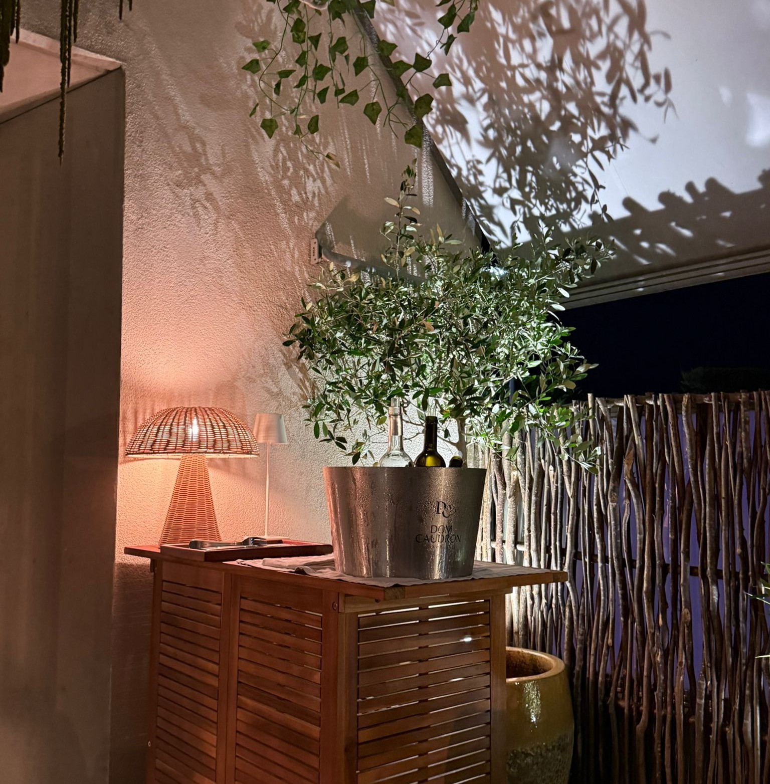 Seau à champagne argenté sur meuble bois à lamelles, lampe osier rose et olivier en pot, terrasse de l'Auberge d'Arthaz la nuit
